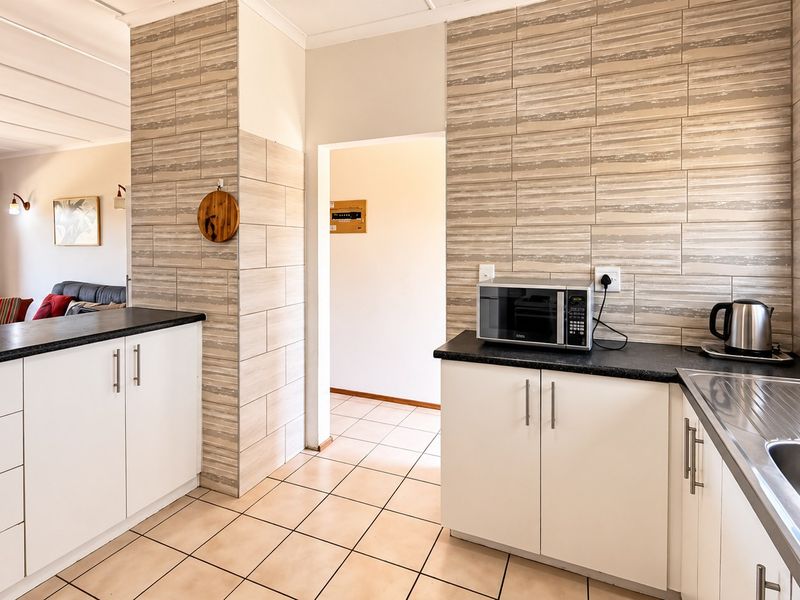 A functional kitchen featuring white cabinetry, dark laminate countertops, tiled splashbacks, and tiled flooring, with an open-plan layout connecting to the living area.