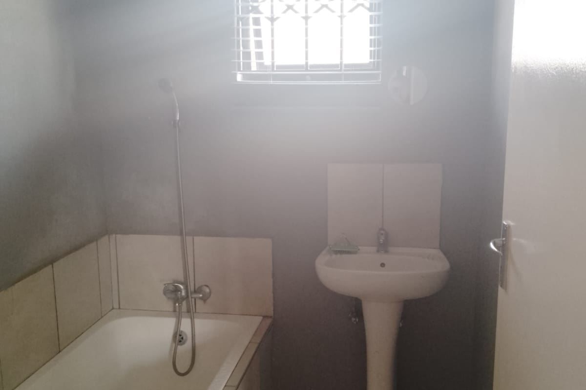 A functional bathroom featuring a pedestal basin, a built-in bathtub with shower mixer, and tiled finishes, illuminated by a window with security bars.