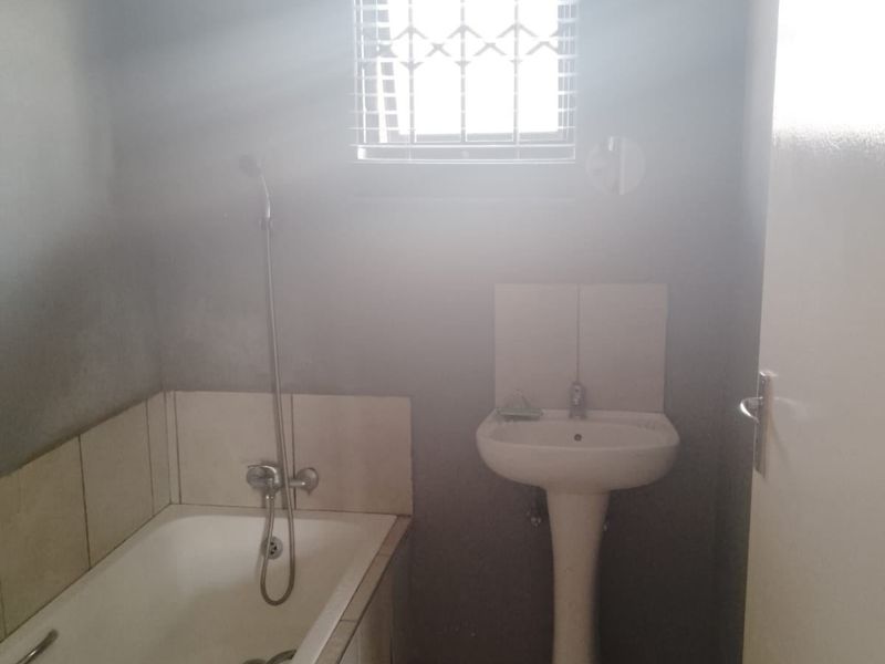 A functional bathroom featuring a pedestal basin, a built-in bathtub with shower mixer, and tiled finishes, illuminated by a window with security bars.
