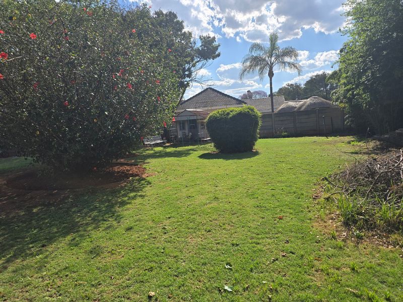 A spacious backyard garden featuring a well-maintained lawn, mature landscaping, and a view of the rear building elevation with a thatched roof.