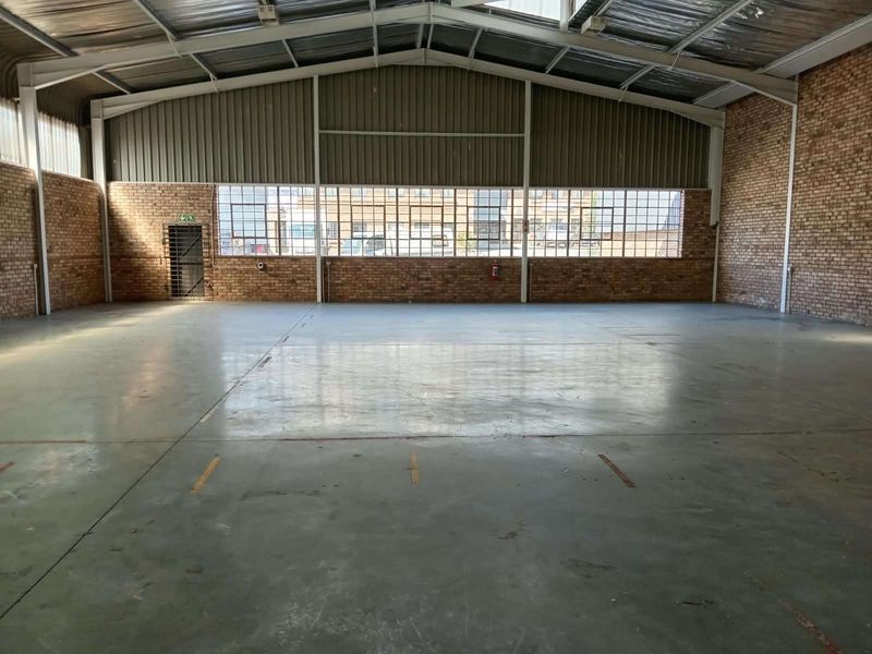 Spacious industrial warehouse unit featuring exposed brick walls, high steel portal frame ceilings, concrete flooring, and large grid windows providing ample natural light.