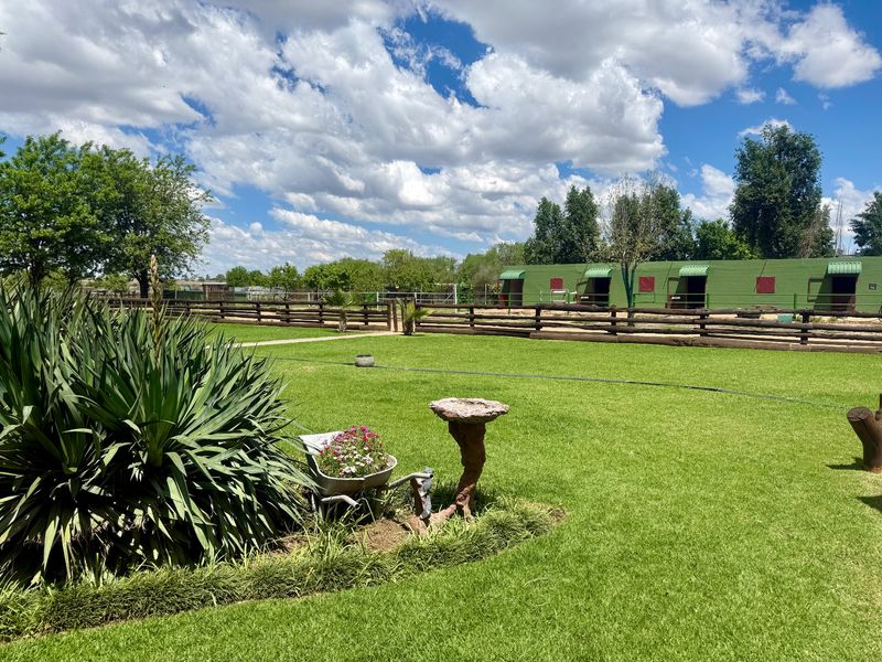 A well-maintained farm garden featuring lush green lawn, timber post and rail fencing, and a row of stables in the background.