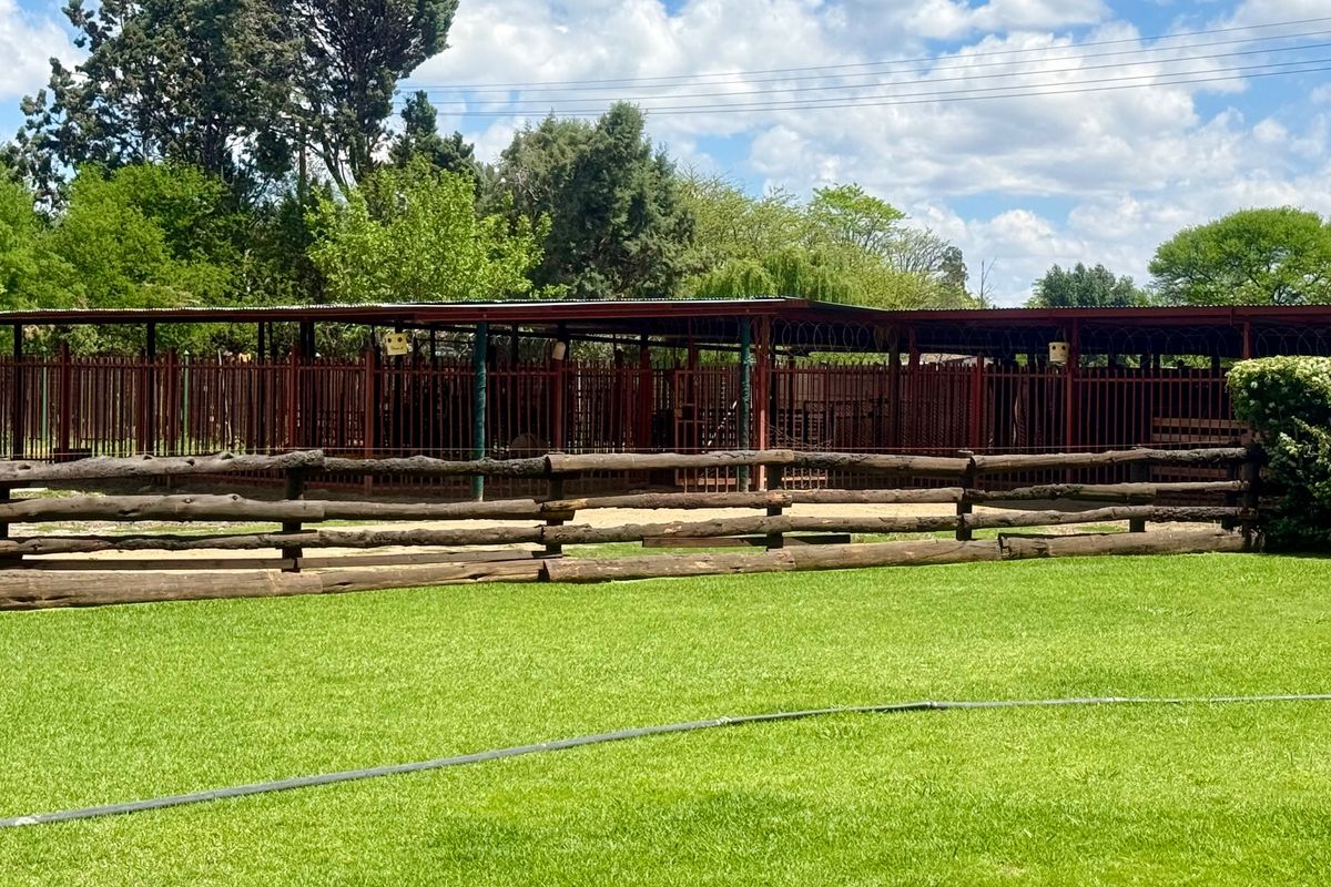 A well-maintained farm area featuring a log fence, metal security fencing, and a covered animal enclosure set against a lush green lawn.
