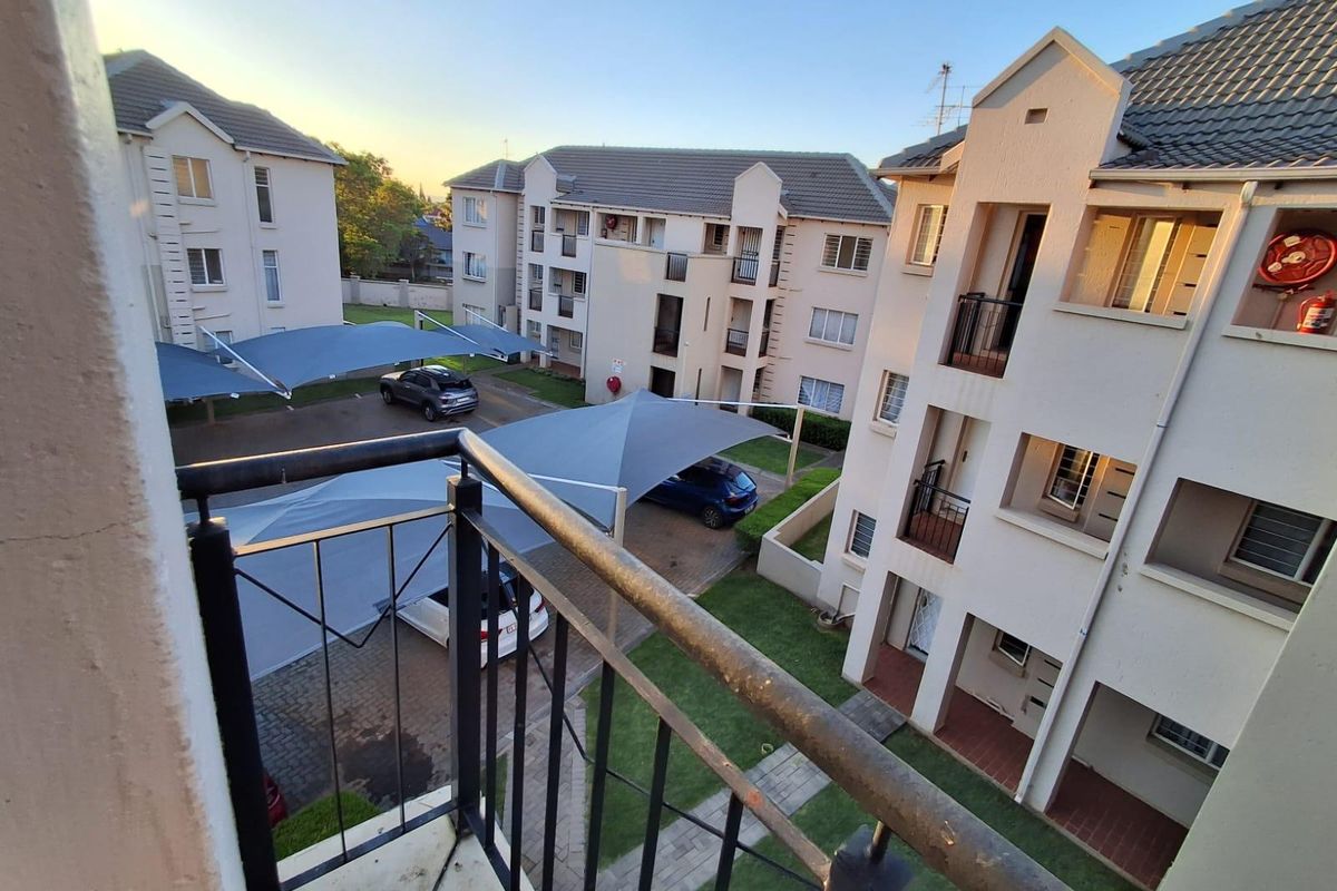 Elevated view from a balcony overlooking the apartment complex grounds, featuring paved parking areas with shade cloth carports and communal garden spaces.