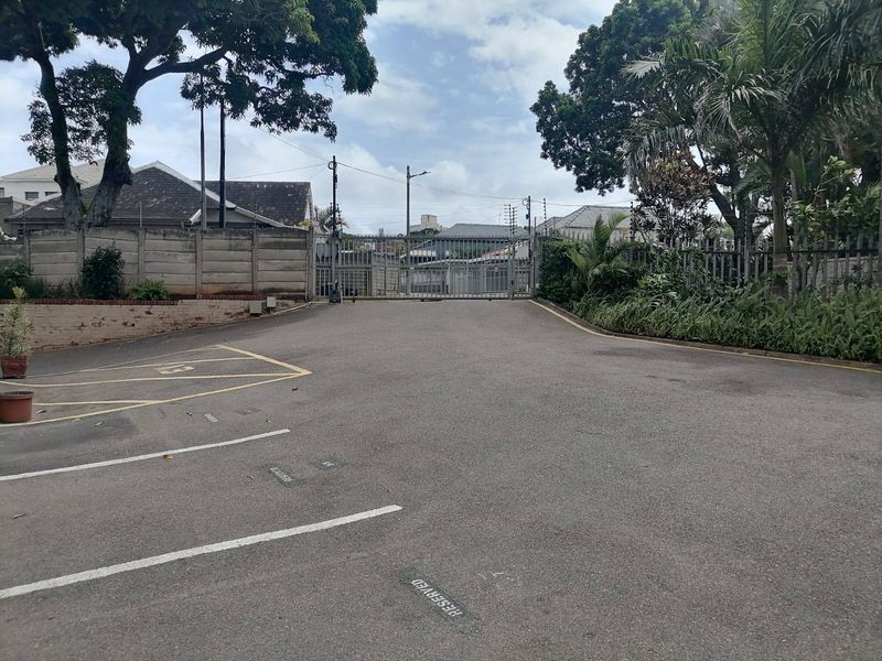 Exterior view of the apartment complex entrance featuring a paved driveway, marked parking bays, and secure gated access with perimeter walling.