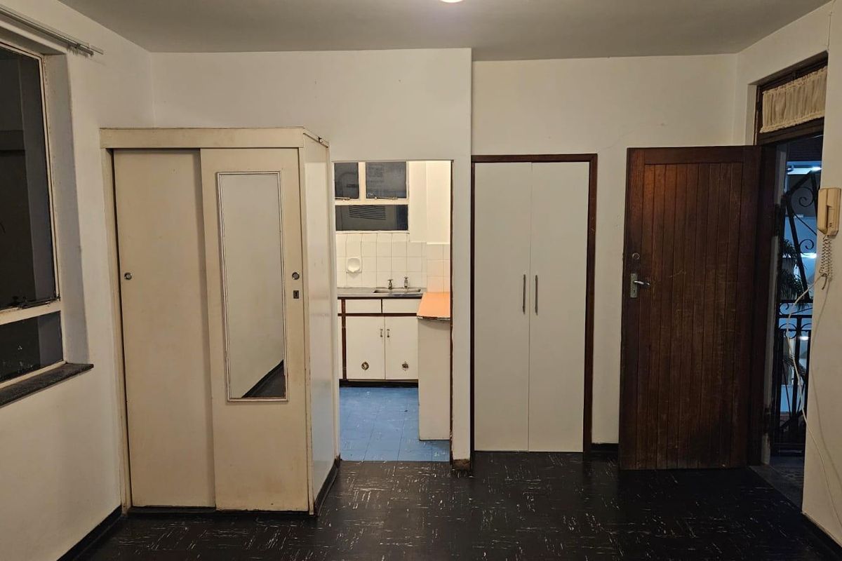 An empty living area featuring built-in cupboards, a timber entrance door, and an intercom system, with a view through to the adjacent kitchen.