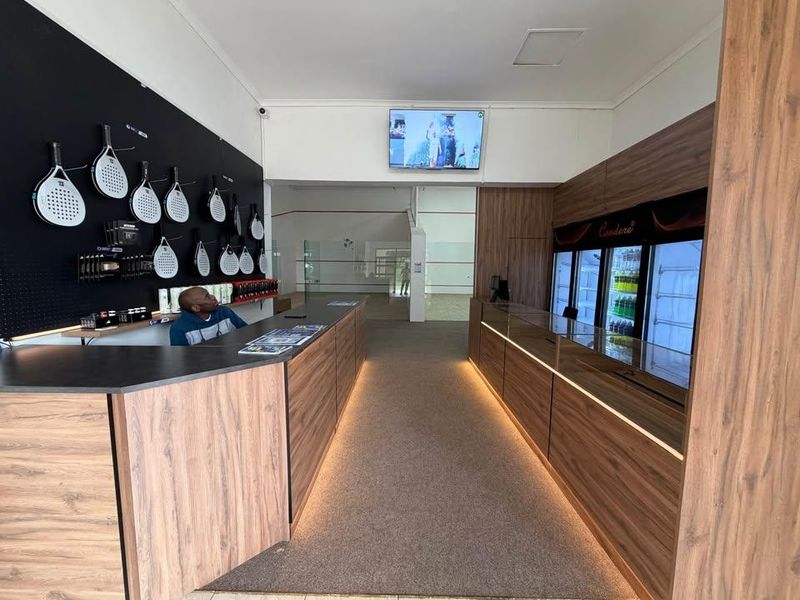 Modern clubhouse reception area featuring wood-grain cabinetry, integrated display fridges, and professional under-counter lighting, leading into a sports facility.