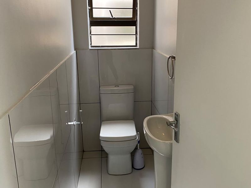 A compact guest bathroom featuring a white pedestal basin, toilet, and neutral wall and floor tiling with a louvered window for ventilation.
