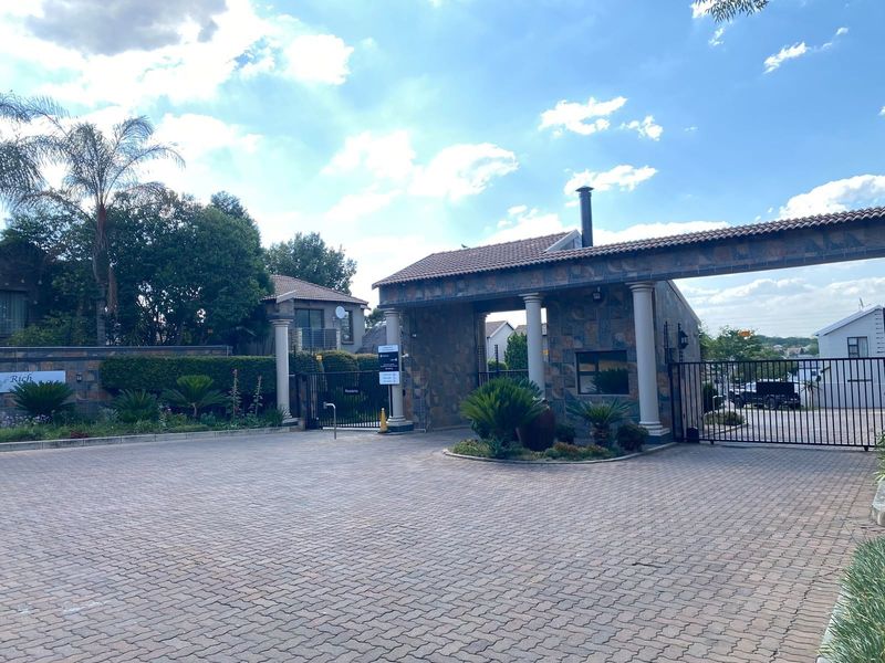 The entrance to the residential complex features a secure gated access point with stone-clad pillars, a guard house, and a well-maintained paved driveway.