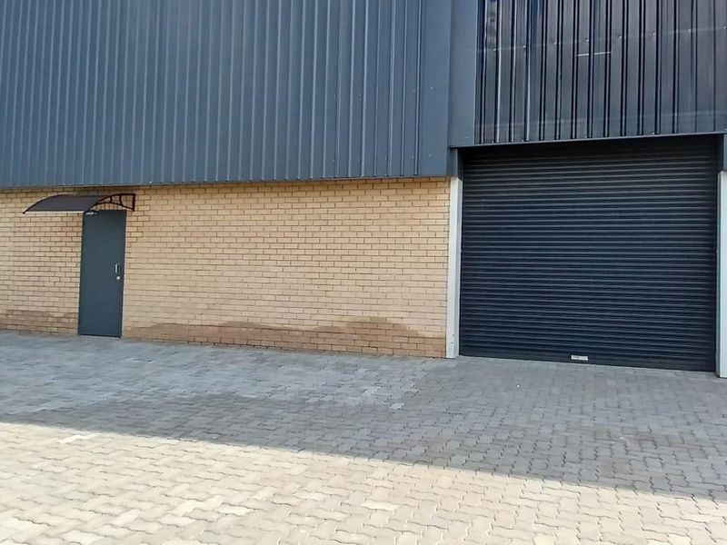 Industrial warehouse exterior featuring a large roller shutter door, pedestrian access points, and paved driveway area with visible brickwork.