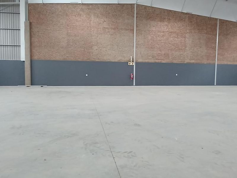 Spacious industrial warehouse interior featuring high ceilings with exposed steel trusses, concrete flooring, and a combination of brick and painted wall finishes.