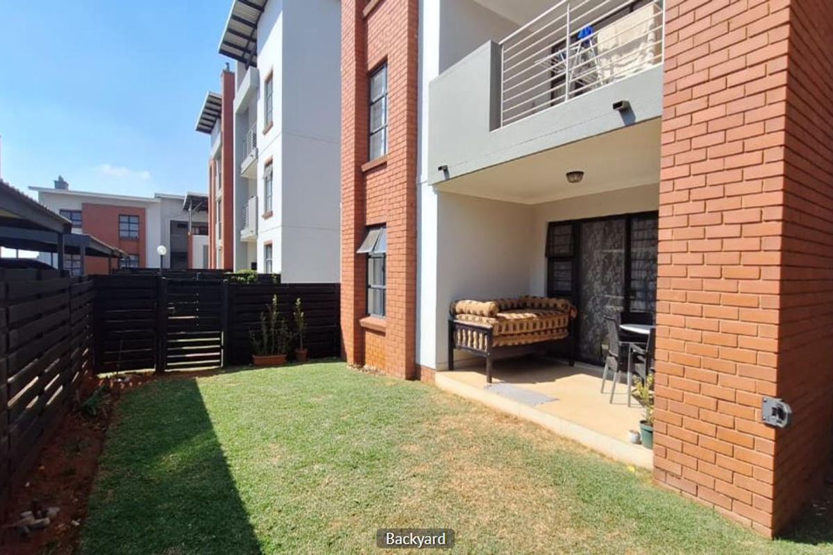 A private rear garden featuring a lawn area, a covered ground-floor patio, and an upper-level balcony with metal railings in a modern apartment complex.