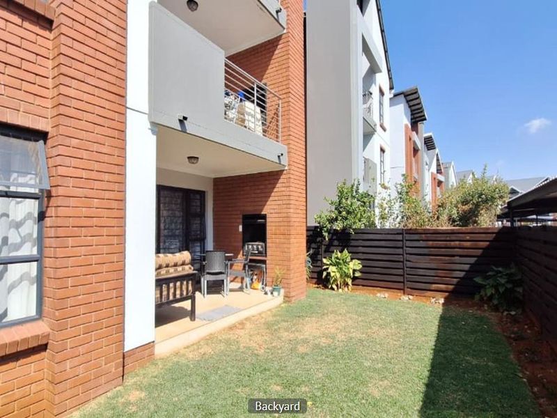 A private rear garden featuring a lawn, timber fencing, and a covered patio area equipped with a built-in braai and overhead balcony.