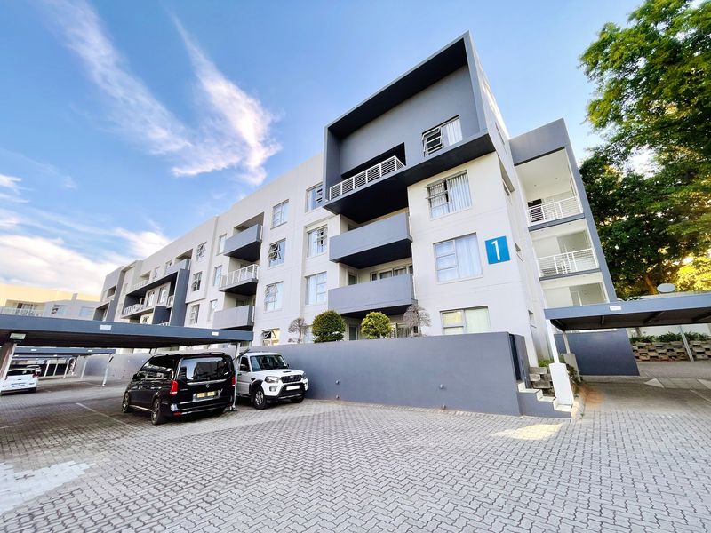 Modern multi-story apartment building exterior featuring a paved driveway, covered parking bays, and private balconies.