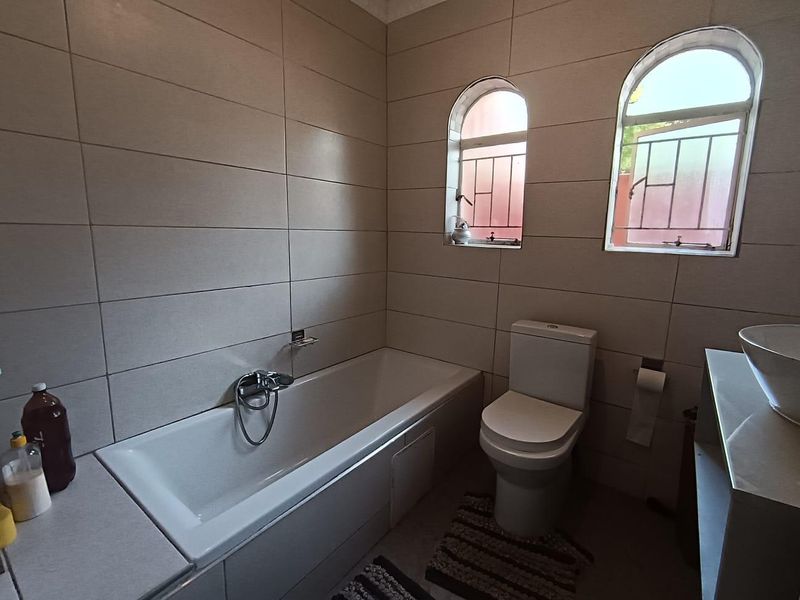 A functional bathroom featuring a bathtub, toilet, and basin, finished with neutral wall tiling and two arched windows with security bars.
