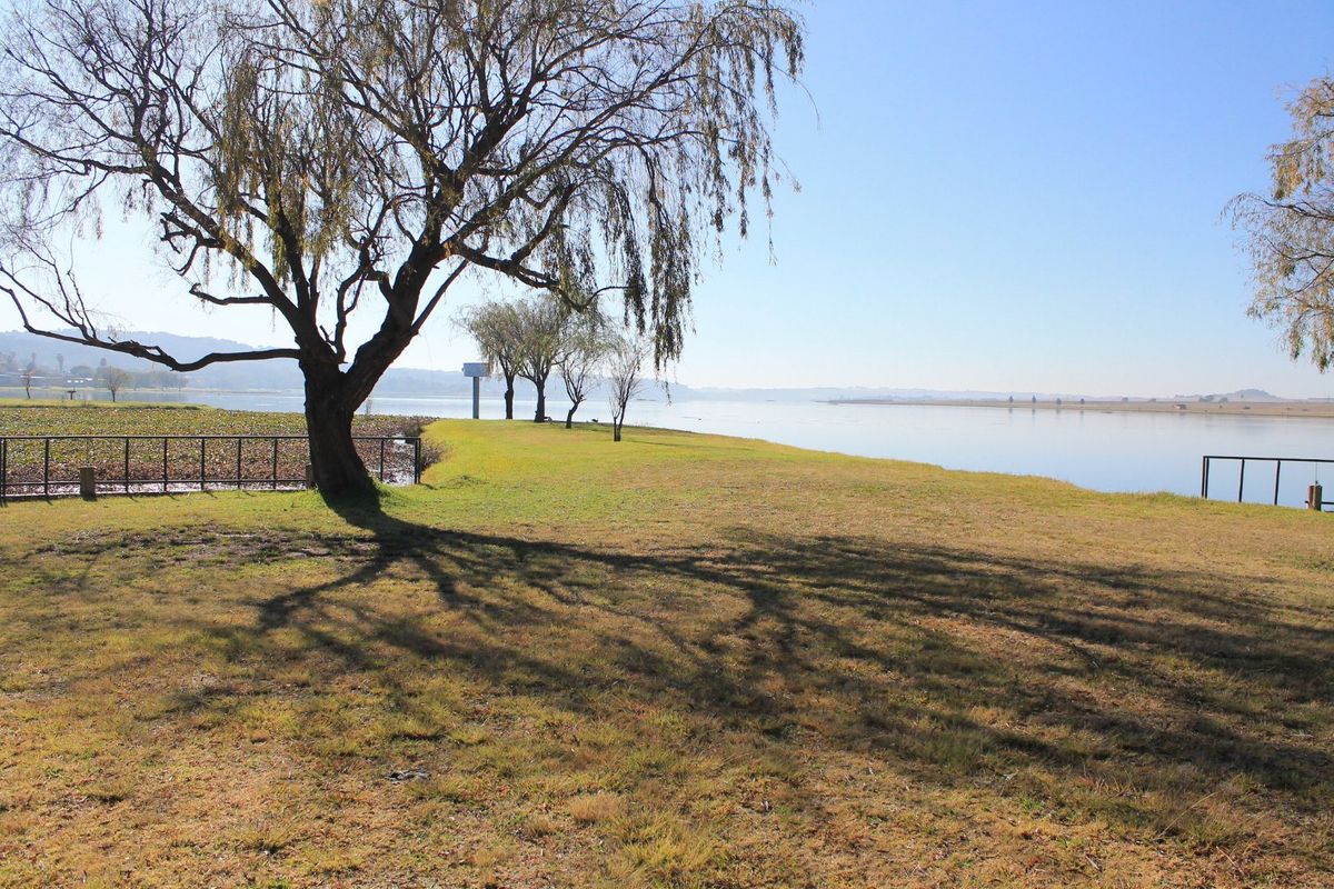 A scenic view of the waterfront property featuring mature trees, open grass areas, and metal perimeter fencing along the water's edge.
