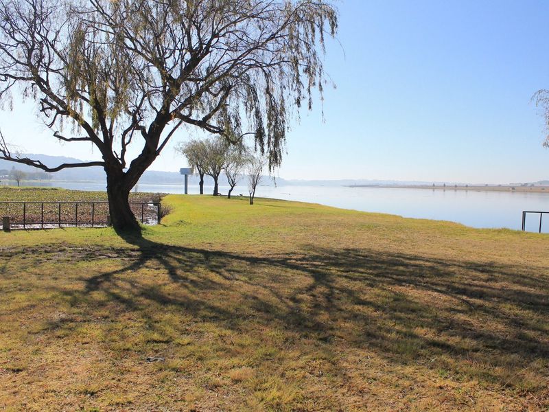 A scenic view of the waterfront property featuring mature trees, open grass areas, and metal perimeter fencing along the water's edge.