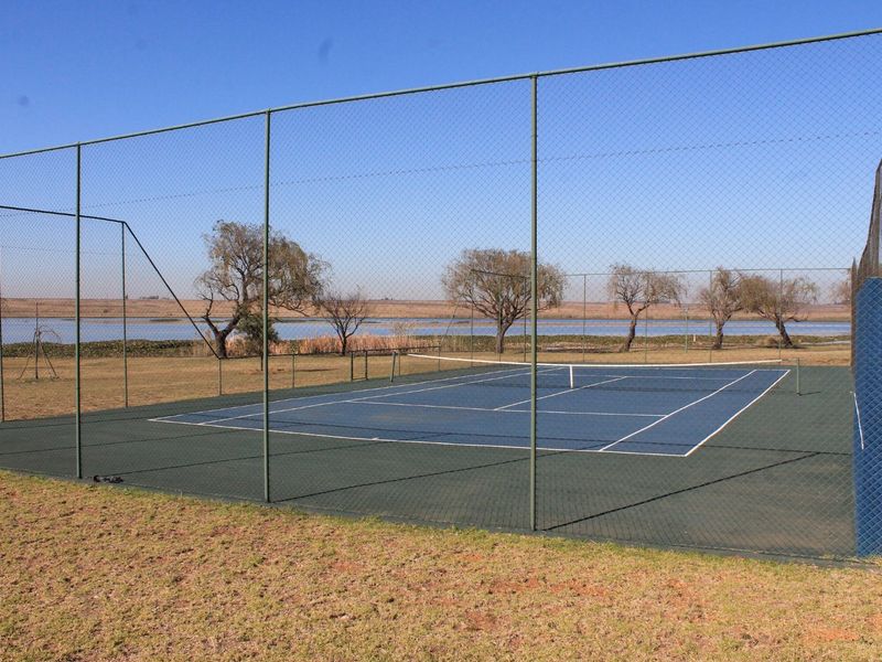 A well-maintained outdoor tennis court enclosed by high chain link fencing, situated in a scenic landscape overlooking a lake.