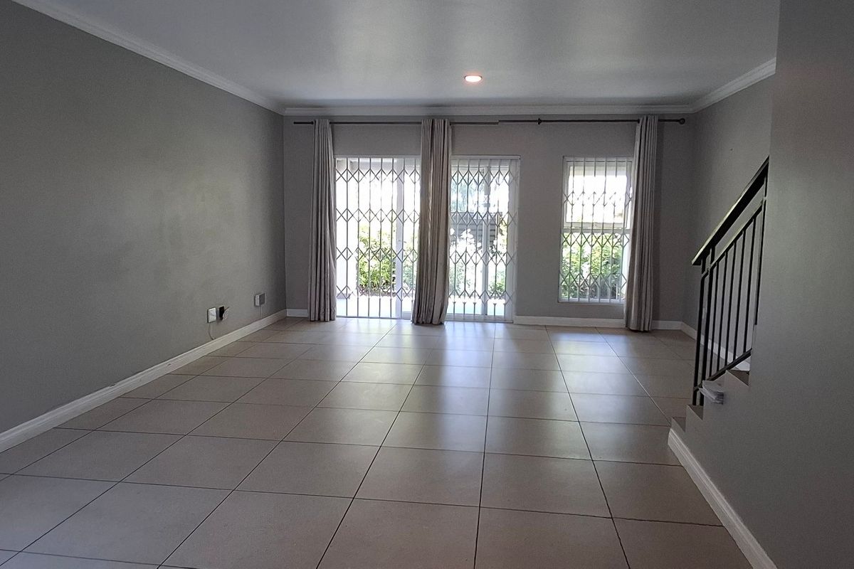 A spacious, empty living area featuring neutral tiled flooring, recessed lighting, and a staircase with a metal railing, leading to a secure patio entrance.
