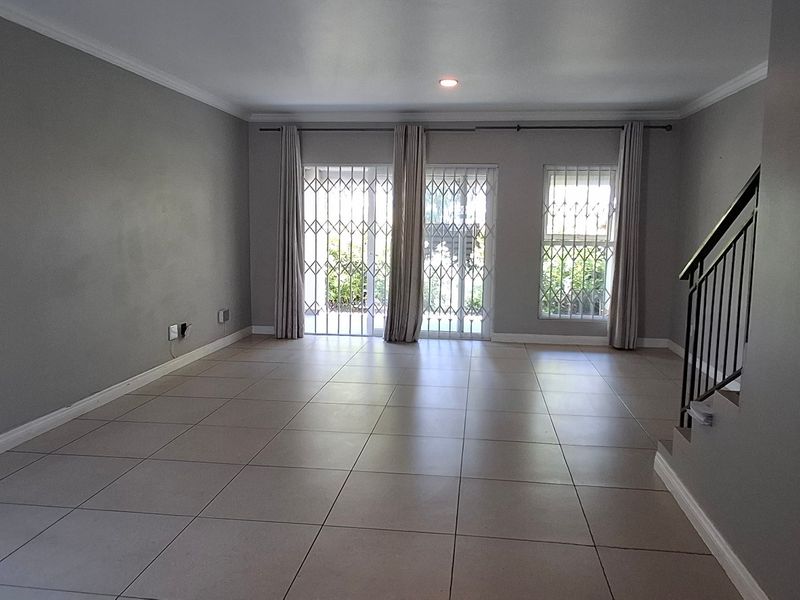 A spacious, empty living area featuring neutral tiled flooring, recessed lighting, and a staircase with a metal railing, leading to a secure patio entrance.
