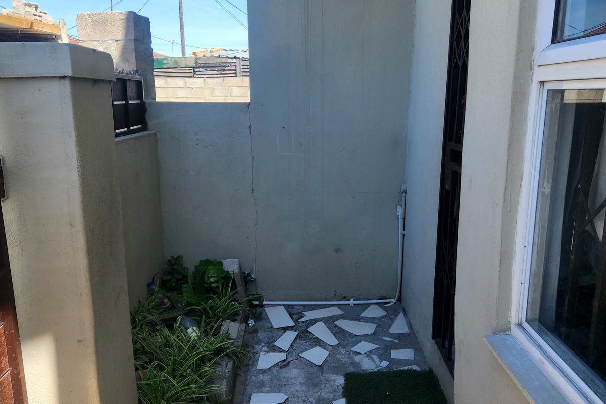 A small rear exterior courtyard area featuring boundary walls, security gate, and window access, currently requiring maintenance to the flooring and wall surfaces.