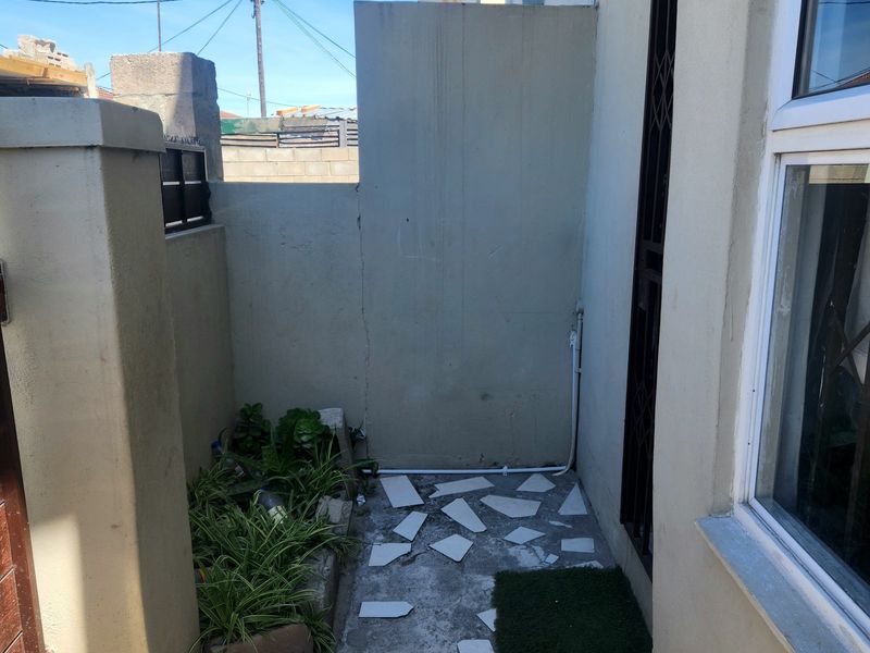 A small rear exterior courtyard area featuring boundary walls, security gate, and window access, currently requiring maintenance to the flooring and wall surfaces.