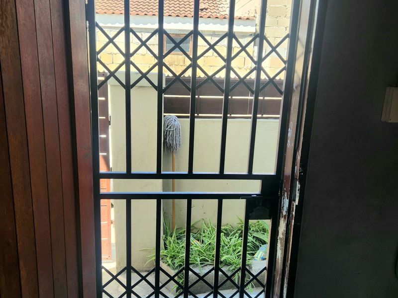 View through a security gate looking out onto a paved rear courtyard area with boundary walls and tiled roof structure.