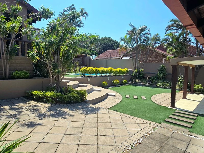 A well-maintained rear garden featuring a paved patio, artificial lawn, covered outdoor entertainment area, and tiered landscaping with retaining walls.