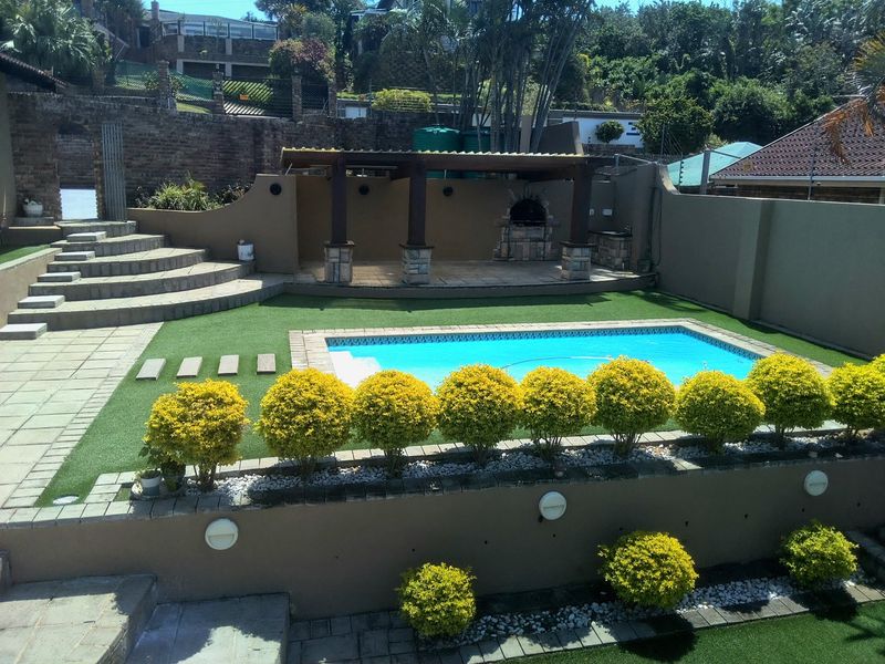 Well-maintained outdoor entertainment area featuring a swimming pool, covered pergola with built-in braai, tiered retaining walls, and landscaped garden beds.