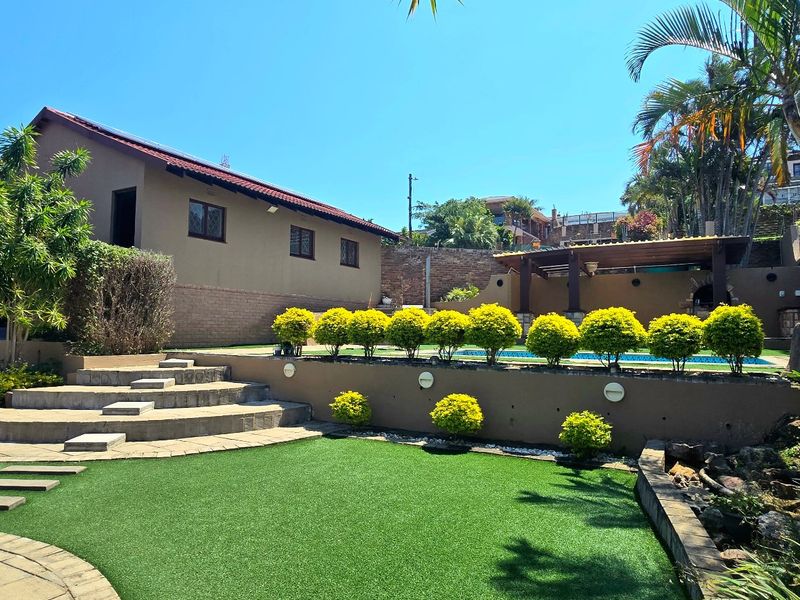 A well-maintained rear garden featuring a swimming pool, tiered retaining walls, paved patio areas, and a built-in braai, perfect for outdoor entertaining.