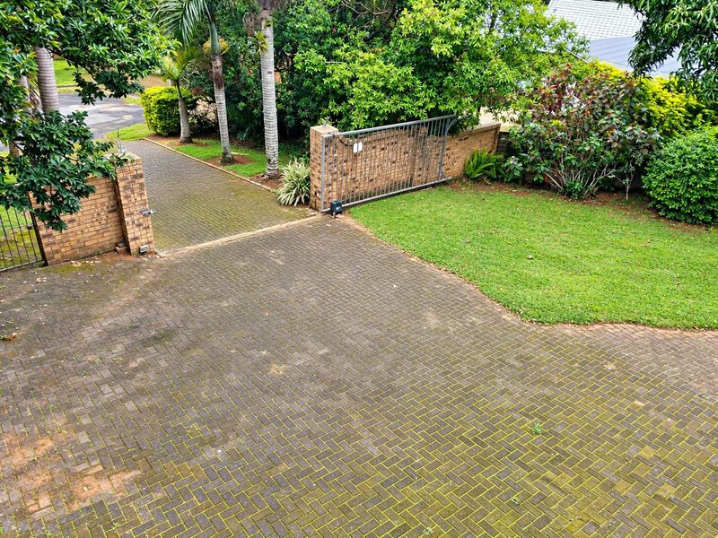 An elevated view of a paved driveway entrance featuring a sliding gate, brick boundary walls, and established garden landscaping.