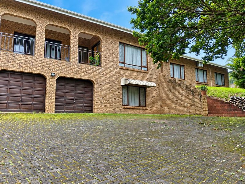A multi-level brick residence featuring a double garage, upper-level balcony with metal railings, and a paved driveway with mature landscaping.