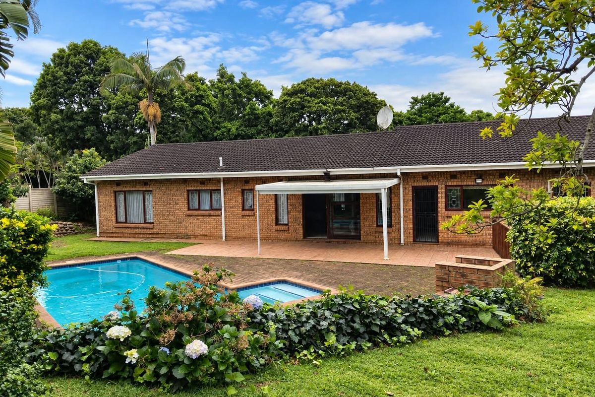 Rear view of a brick house featuring a swimming pool, paved patio area, and a built-in braai, set within a landscaped garden.