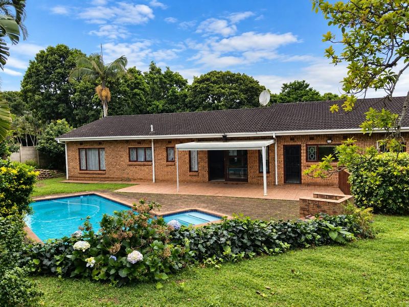 Rear view of a brick house featuring a swimming pool, paved patio area, and a built-in braai, set within a landscaped garden.
