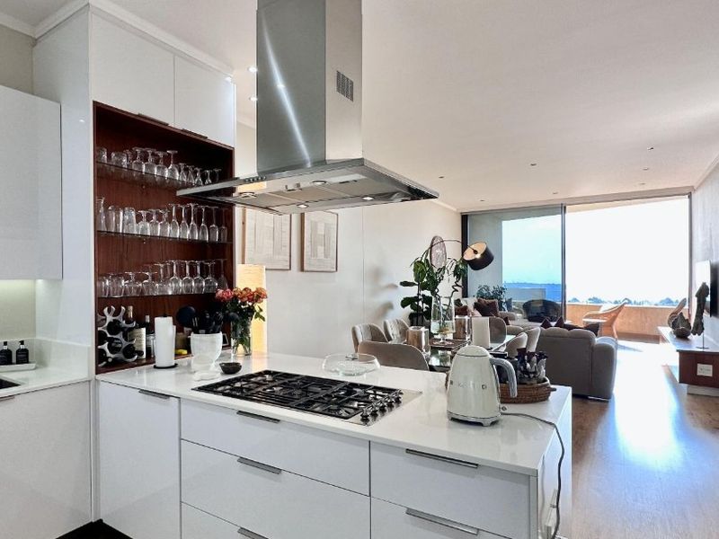 Modern open-plan kitchen featuring sleek white cabinetry, stone countertops, and a gas hob, seamlessly flowing into the living area with expansive balcony access.