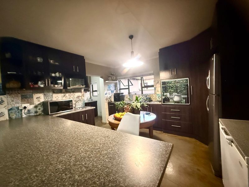 A functional kitchen featuring dark wood cabinetry, granite-look laminate countertops, and a decorative tile backsplash, with an open-plan layout connecting to the dining area.