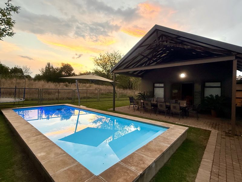 Outdoor entertainment area featuring a swimming pool with tiled coping and a covered patio with exposed roof trusses, set against a sunset backdrop.