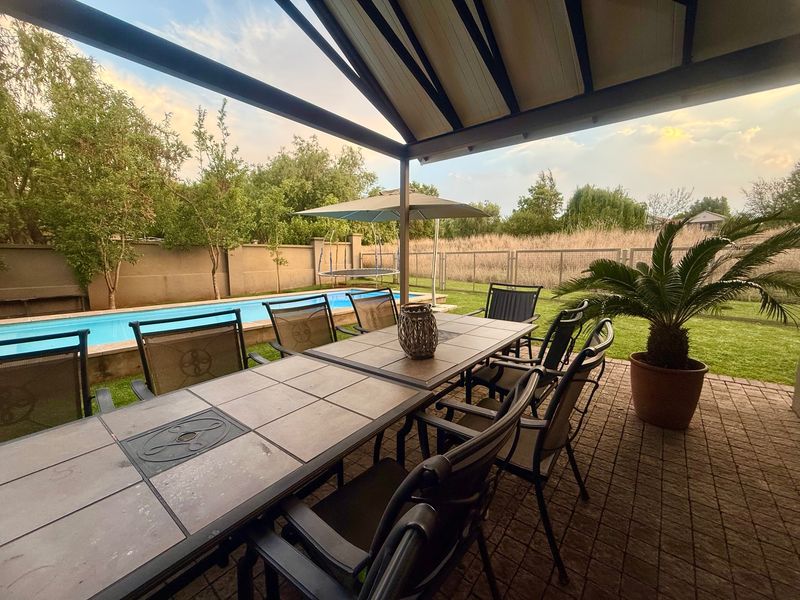 A covered outdoor patio area overlooking a swimming pool and garden, featuring paved flooring and a structural roof covering.