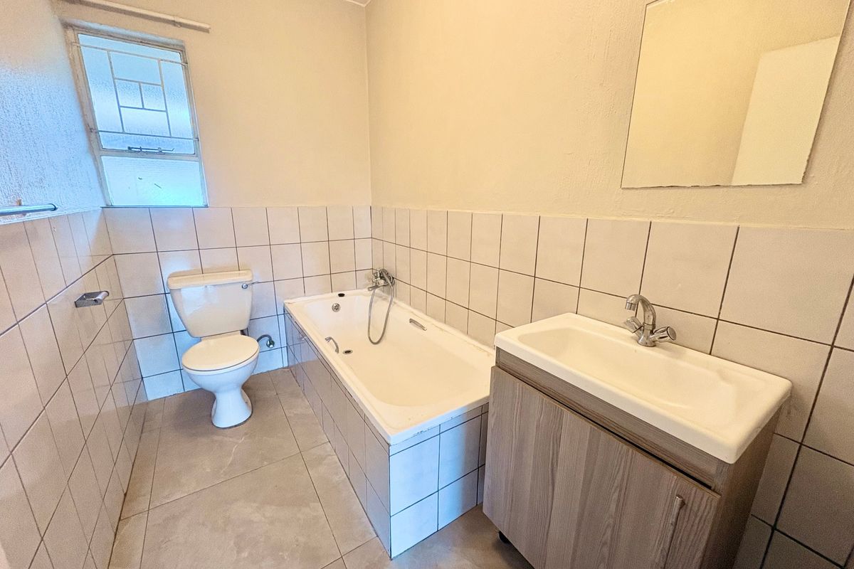 A functional bathroom featuring a built-in bathtub, vanity cabinet with basin, and toilet, finished with neutral wall and floor tiling.