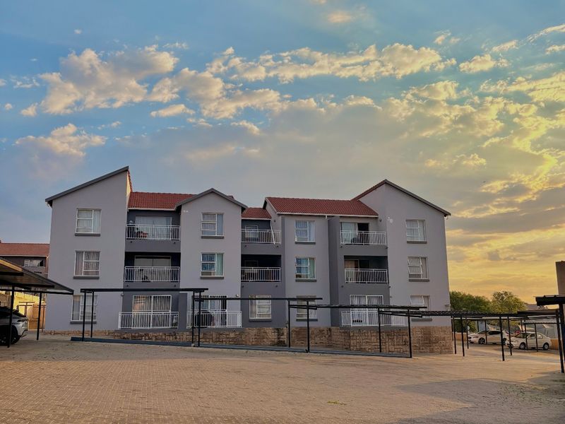A multi-story apartment building featuring a tiled roof, private balconies, and a paved parking area with carports.