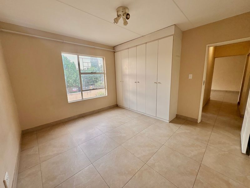 A vacant bedroom featuring neutral tiled flooring, built-in storage cupboards, and a window with security bars.