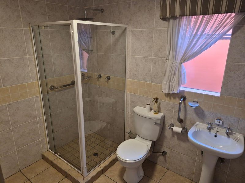 A functional bathroom featuring a glass-enclosed shower, pedestal basin, and toilet, finished with neutral wall and floor tiling and equipped with safety grab rails.