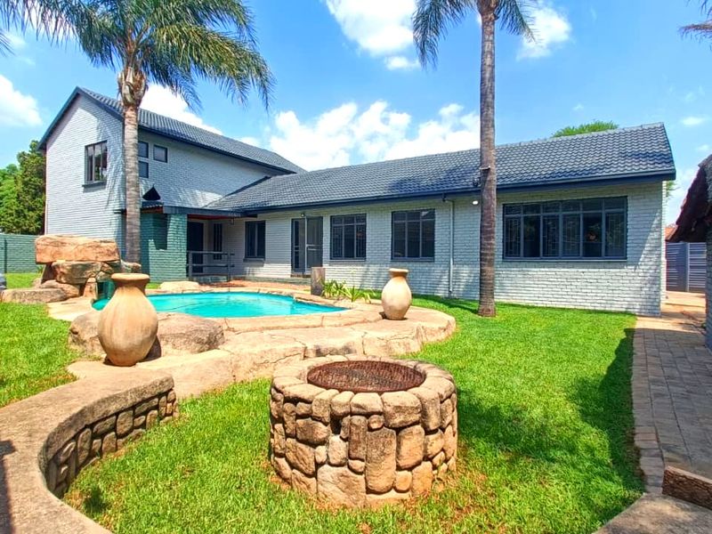Rear exterior view featuring a swimming pool, stone fire pit, landscaped garden, and a thatched lapa area, perfect for outdoor entertaining.