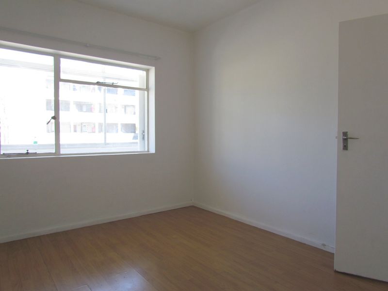 A bright, empty bedroom featuring laminate flooring, a large sliding window, and a neutral white interior finish.