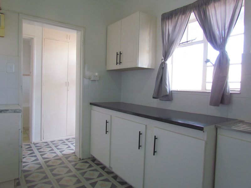 A functional kitchen featuring white built-in cabinetry, dark laminate countertops, tiled splashbacks, and patterned floor tiles, leading into a hallway with built-in storage.