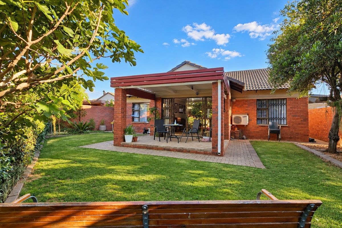 Rear view of a townhouse featuring a brick facade, a covered patio area, and a well-maintained garden with mature trees.