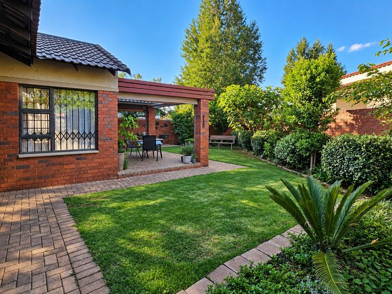 A well-maintained rear garden featuring a paved patio area, lush lawn, and established landscaping, enclosed by a brick boundary wall.