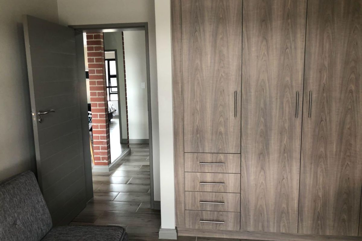 A bedroom featuring modern wood-grain built-in cupboards, tiled flooring, and a view through to an adjacent room with exposed brick detailing.