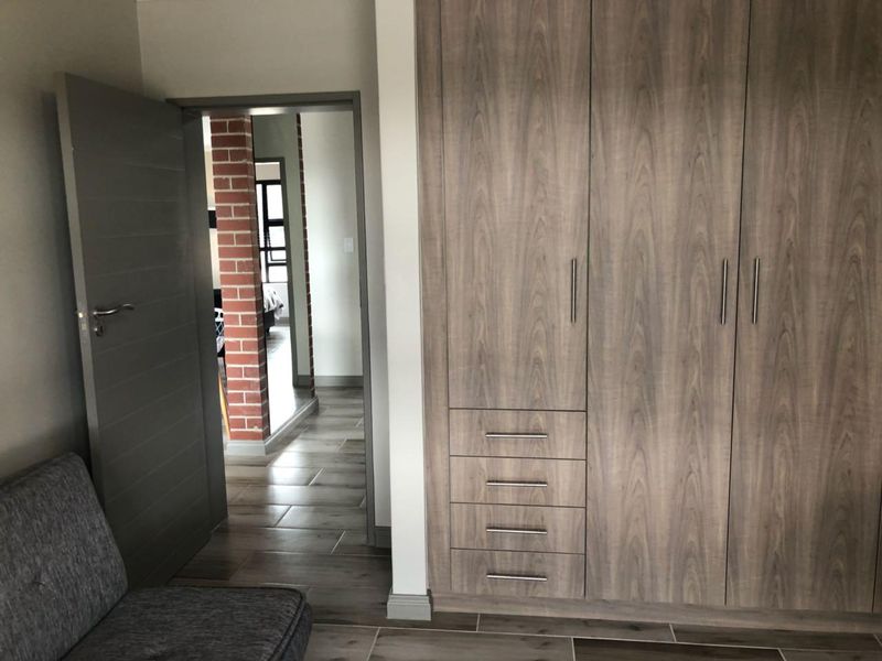 A bedroom featuring modern wood-grain built-in cupboards, tiled flooring, and a view through to an adjacent room with exposed brick detailing.