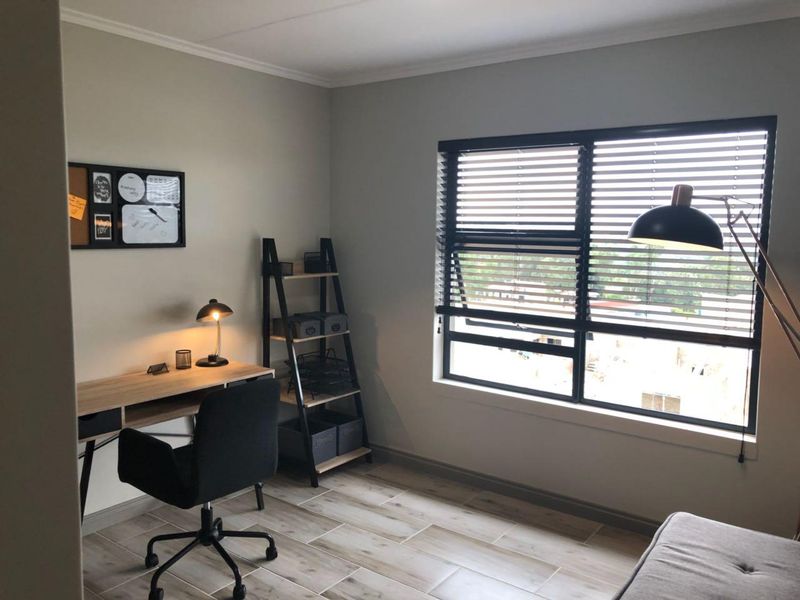 A well-lit home office featuring modern tiled flooring, large aluminium-framed windows with venetian blinds, and neutral wall finishes.